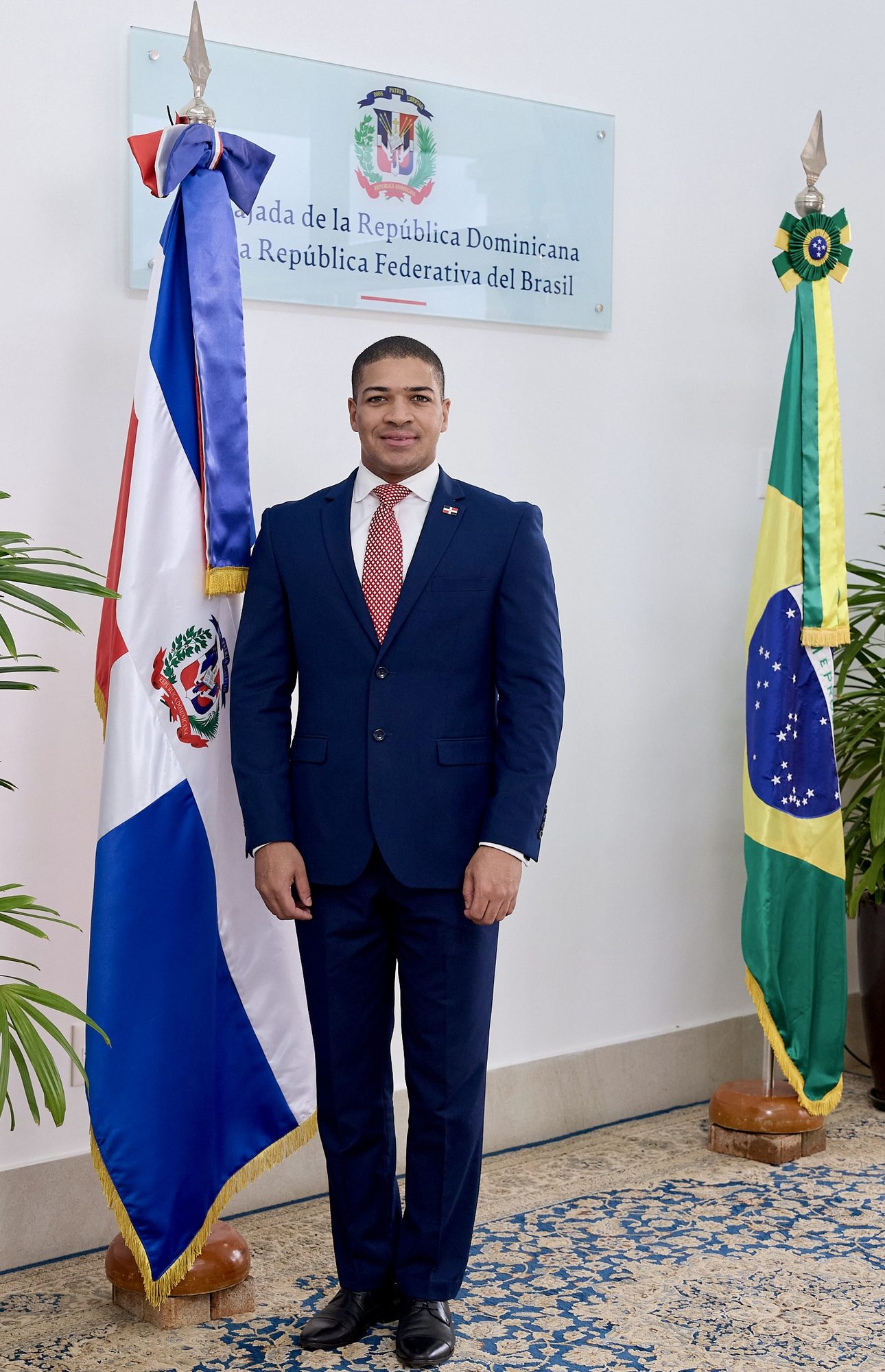 Jovem diplomata entre as bandeiras da República Dominicana e do Brasil sob a placa da embaixada