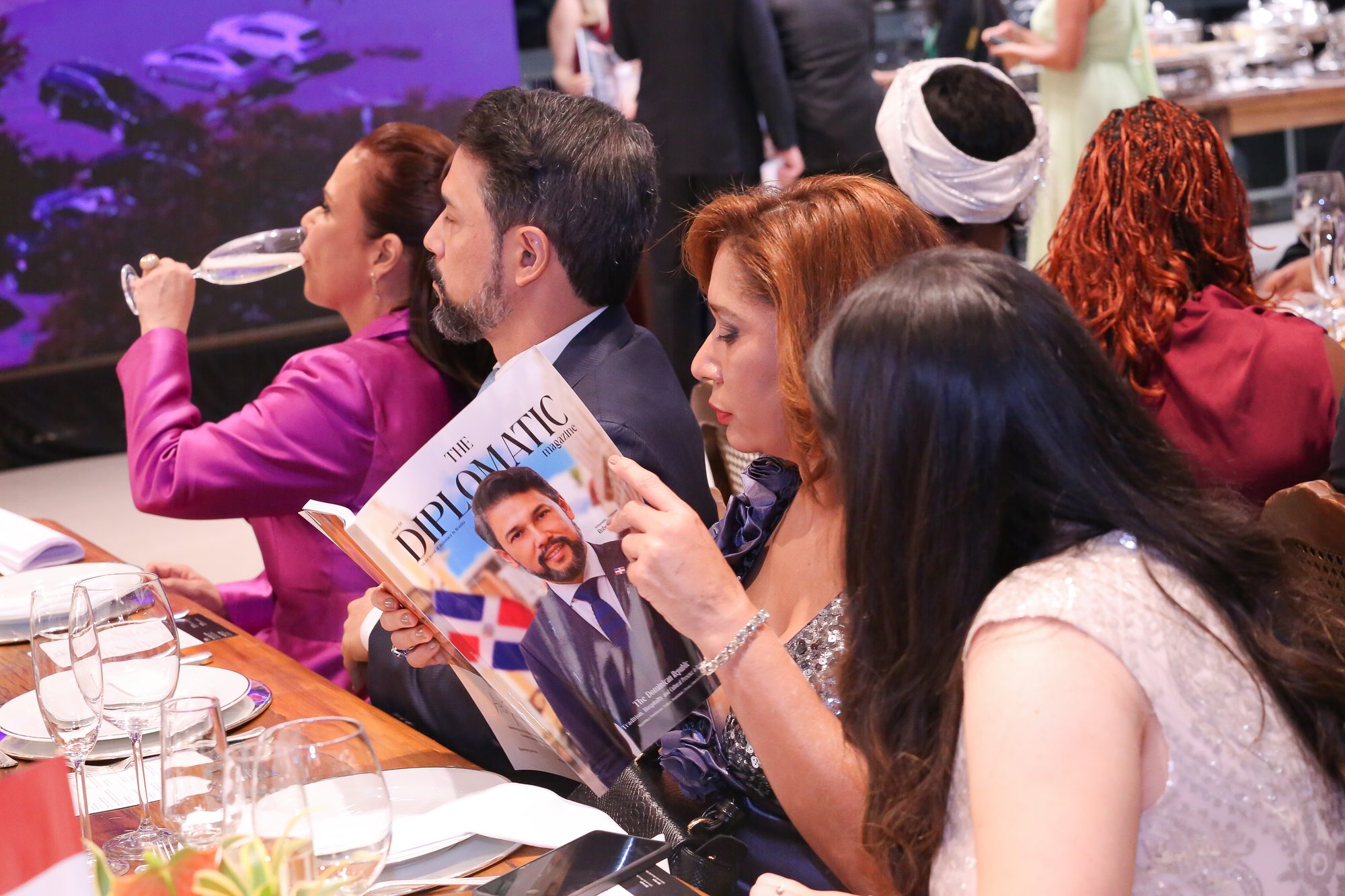 Convidados folheando a revista The Diplomatic à mesa de gala, ao lado da bandeira dominicana